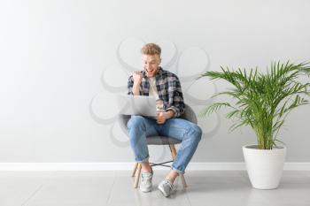 Happy young man with laptop sitting on chair near light wall�