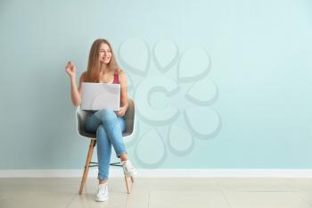 Beautiful young woman with laptop sitting on chair against color wall�