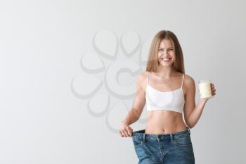 Beautiful young woman in loose jeans and with jar of coconut oil on light background. Weight loss concept�