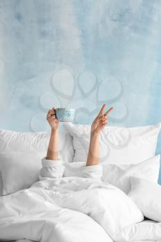 Young woman with cup of hot beverage showing victory gesture in bed�