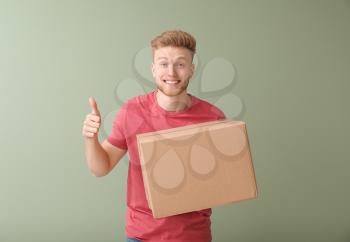 Young man with cardboard box showing thumb-up on color background�