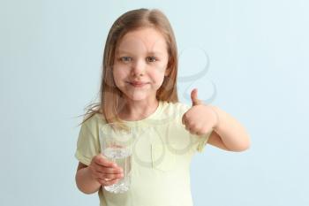 Cute little girl with glass of water showing thumb-up gesture on light background�