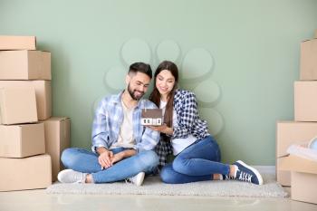 Young couple with belongings and model of house sitting near color wall�