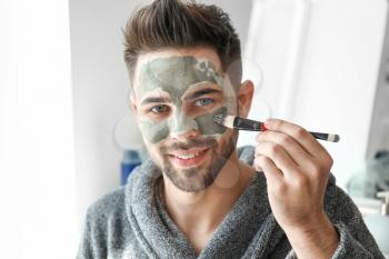 Handsome man applying clay mask onto his face at home�