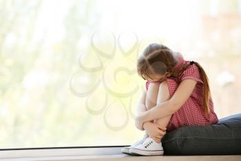 Sad little girl with autistic disorder sitting on window sill at home�
