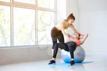Young sporty woman with fitball doing exercises under supervision of her personal trainer in gym�