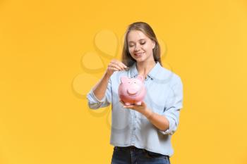Young woman putting coin into piggy bank on color background�
