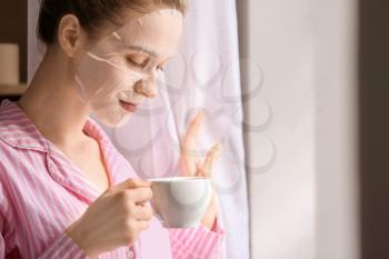 Young woman with sheet facial mask drinking tea at home�