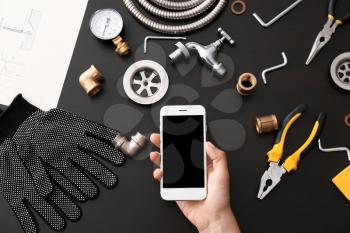 Plumber tools and female hand with mobile phone on dark background, top view�
