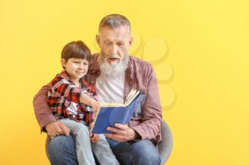 Cute little boy reading book with grandfather on color background�