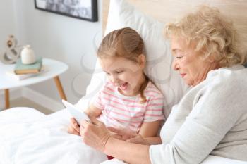 Cute little girl with grandmother using tablet PC in bed at home�