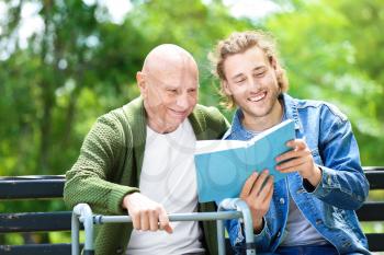 Young man reading book to his elderly father in park�