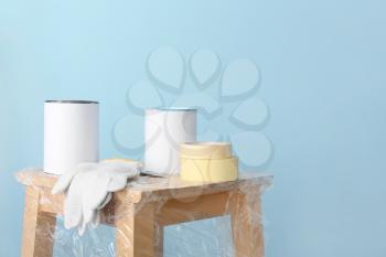 Cans of paint with gloves and tape on stool against color background�