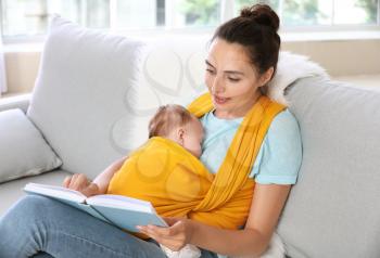Young mother with little baby in sling reading book at home�