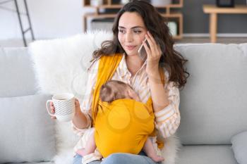 Young mother with little baby in sling talking by mobile phone at home�