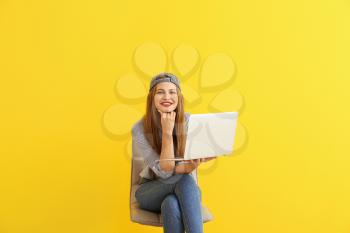 Happy young woman with laptop sitting on chair against color background�