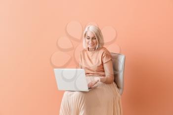 Mature woman with laptop sitting on chair against color background�