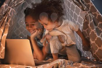 Cute African-American sisters with laptop watching cartoons in hovel�