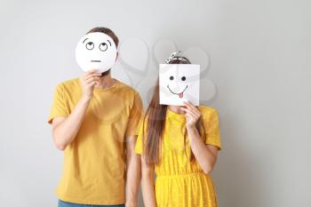 Couple hiding faces behind sheets of paper with drawn emoticons on grey background�