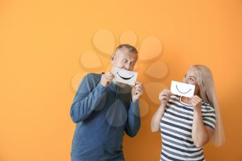 Mature couple holding paper sheets with drawn smiles against color background�
