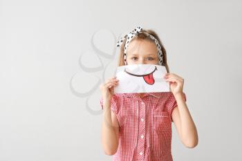 Little girl hiding mouth behind drawn smile on light background�