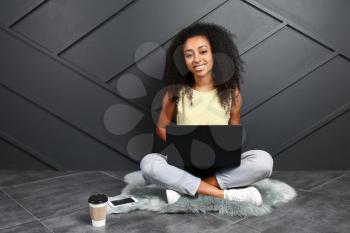 Portrait of beautiful African-American woman with laptop sitting near dark wall�