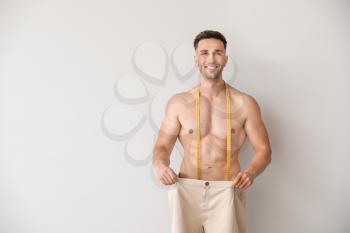 Handsome muscular man in loose pants and with measuring tape on light background. Weight loss concept�