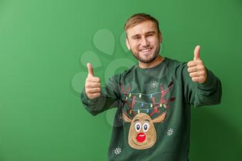 Young man in Christmas sweater showing thumb-up on color background�