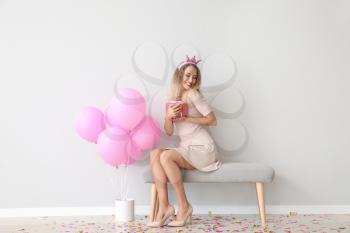 Beautiful young woman with balloons and gift sitting near light wall�