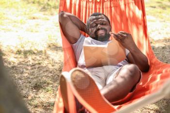African-American man reading book while relaxing in hammock outdoors�