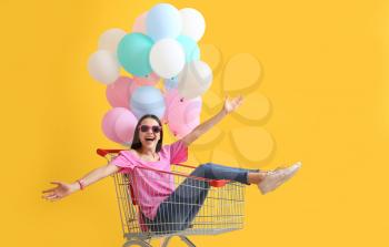 Young woman with shopping cart and balloons on color background�