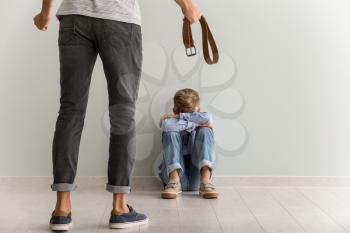 Man threatening his little son who is sitting on floor near light wall. Physical punishment concept�
