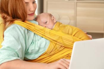 Young mother with little baby in sling using laptop at home�