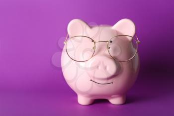 Piggy bank with eyeglasses on color background�