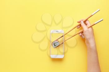Female hand holding chopsticks and mobile phone with tasty sushi roll on screen against color background�