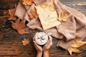 Female hands with hot chocolate in cup, warm sweater and dry leaves on wooden background�