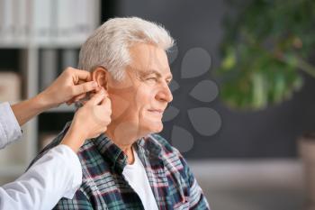 Doctor putting hearing aid in mature man's ear in clinic�