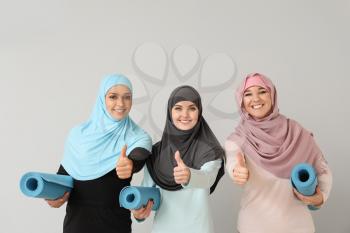 Sporty Muslim women with yoga mats showing thumb-up on light background�