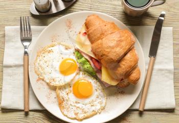 Plate with tasty croissant sandwich and fried eggs on wooden table�