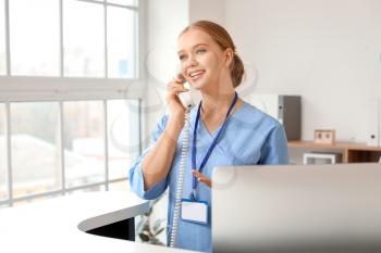 Young female receptionist talking by telephone at desk in clinic�