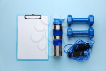 Dumbbells with clipboard, jumping rope and bottle of water on color background�