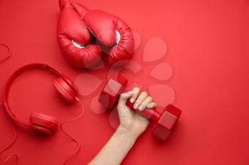 Female hand with dumbbell, boxing gloves and headphones on color background�