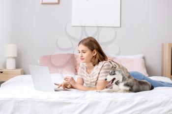 Cute teenage girl with funny husky puppy and laptop on bed at home�