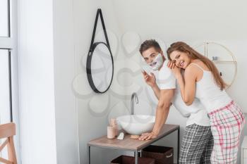 Morning of beautiful young woman and her husband with shaving foam on his face in bathroom�