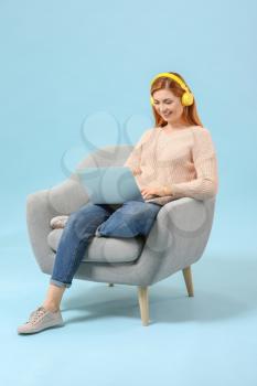 Beautiful woman with laptop listening to music while sitting in armchair against color background�