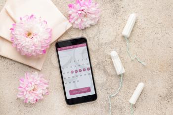 Menstrual calendar on screen of mobile phone and feminine products on grey background�