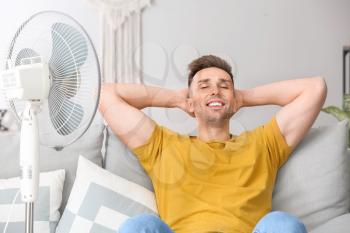 Young man using electric fan during heatwave at home�