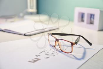 Glasses with eye test chart on table in ophthalmology clinic�