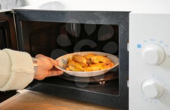 Woman putting plate with fried potato in microwave oven�