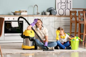 Tired woman and her little daughter with vacuum cleaner in kitchen�